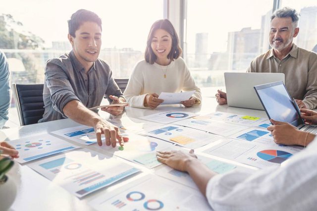 A group of people in a bright office setting are sitting around a table covered with charts and documents. They are discussing and collaborating, with one person using a tablet and another using a laptop. Large windows provide a view of city buildings.