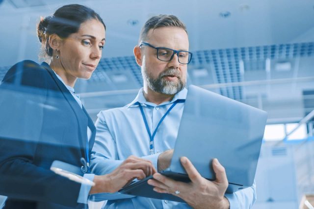 A man and a woman in business attire are standing together, focused on a laptop. The woman has dark hair tied back, and the man has a beard and is wearing glasses. They appear to be in a modern office setting with a ceiling grid and bright lighting.