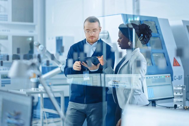 Two people in a modern laboratory setting are discussing something on a tablet. The man wears glasses and a navy cardigan, and the woman wears a light blazer. Advanced equipment and machinery are visible in the background.