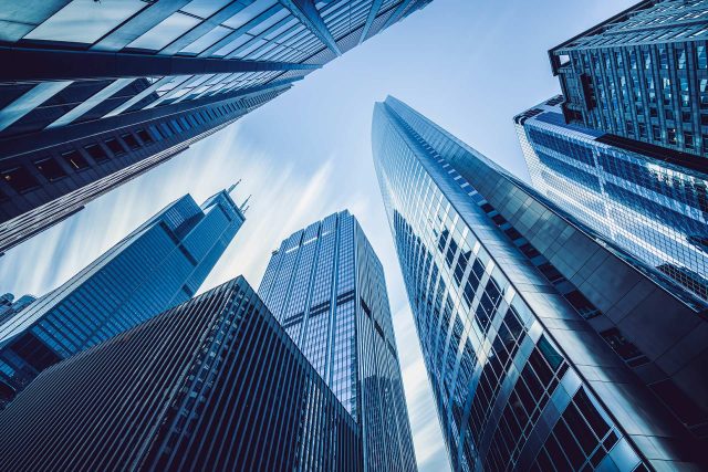 A dramatic upward view of skyscrapers against a clear blue sky, showcasing modern architecture with glass and steel facades. The buildings converge towards the center, creating a dynamic and towering perspective.